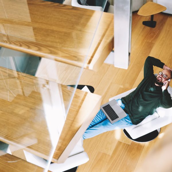 From above of male with toothy smile and hand on head sitting in armchair with laptop on lap having conversation on mobile phone at home