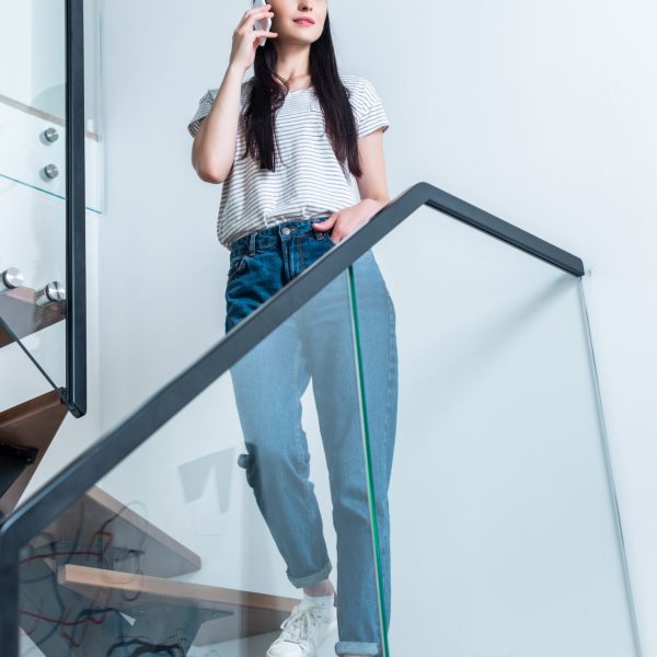 low angle view of attractive woman talking on smartphone while standing on stairs at home