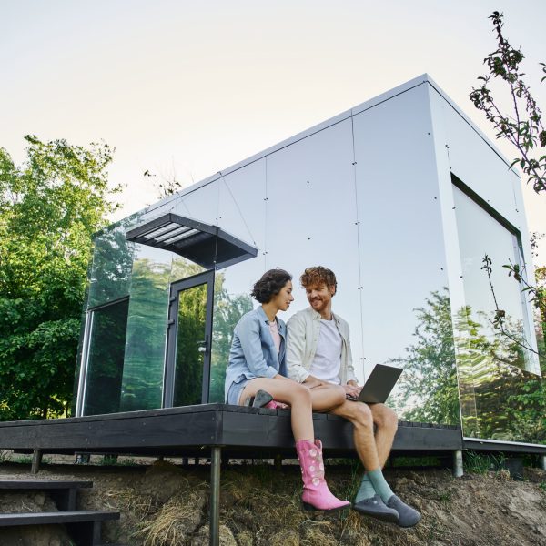 young multiethnic couple sitting on porch with laptop near modern glass house in countryside